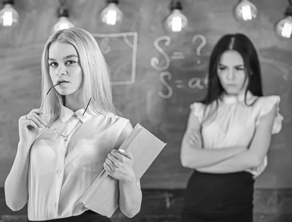 Envy and competition concept. Woman with book and eyeglasses looks dreamy while student on background looks jealous. Girl jealous of success of classmate in classroom, chalkboard on background — Stock Photo, Image