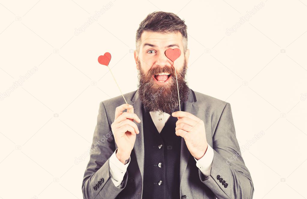Hipster wears smart suit and bow-tie. Mature man with long beard and cheerful face. Bearded man smiles and holds red hearts. Dating, love symbol, happy, fashion, style, celebration, holiday concept