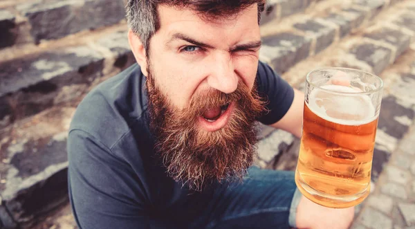 Concepto de celebración. El hombre con barba y bigote sostiene el vaso con cerveza mientras se sienta en escaleras de piedra, desenfocado. Hipster en la cara alegre guiño bebe cerveza al aire libre. Chico descansando con cerveza de barril — Foto de Stock