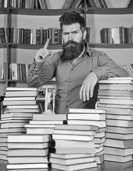 Profesor o estudiante con barba está en la mesa con libros, desenfocado. Concepto bibliotecario. Hombre en rostro seguro se encuentra entre montones de libros, mientras estudia en la biblioteca, estanterías en el fondo —  Fotos de Stock