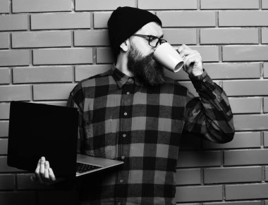 Sakallı, beyaz tenli hippi elinde bardakla dizüstü bilgisayar tutuyor.