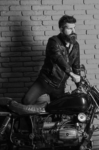 Hipster, motard brutal sur le visage sérieux dans la veste en cuir obtient sur la moto. Concept de passion masculine. Homme avec barbe, motard en veste en cuir près de la moto dans le garage, fond de mur de briques — Photo