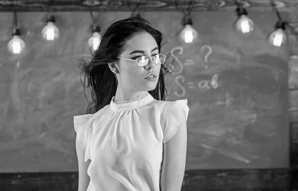 Woman with long hair in white blouse stands in classroom. Lady strict teacher on relaxed face stands in front of chalkboard. Teacher with glasses and waving hair looks sexy. Sexy teacher concept