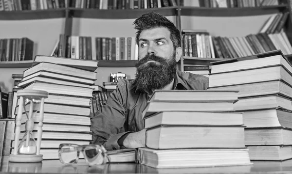 Bookworm concept. Man on strict face sit between piles of books, while studying in library, bookshelves on background. Teacher or student with beard sit at table with hourglass and glasses, defocused — Stock Photo, Image