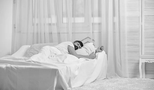 Guy on calm face sleeping on white sheets, pillow. Nap and siesta concept. Macho with beard sleeping, relaxing, having nap, rest. Man laying on bed, covered with blanket, white curtains on background