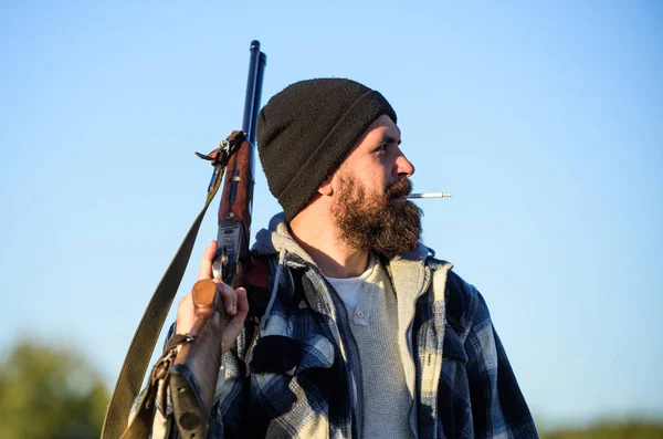 Man brutal bearded guy gamekeeper blue sky background. Hunter with rifle gun close up. Guy bearded hunter spend leisure hunting and smoking. Brutality and masculinity. Hunting masculine hobby concept — Stock Photo, Image