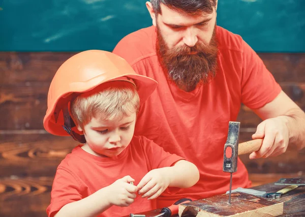 Отец, отец с бородой учит маленького сына пользоваться ногтями и молотком. Мальчик, ребенок занят в защитном шлеме, учится забивать ногти вместе с папой. Концепция мужского долга — стоковое фото