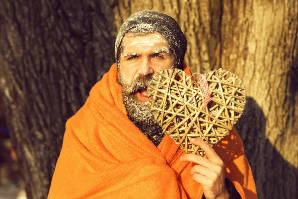 Excited frozen man, bearded hipster, with beard and moustache covered with white frost wrapped in orange blanket with wicker heart on winter day outdoors on natural backgroun — Stock Photo, Image