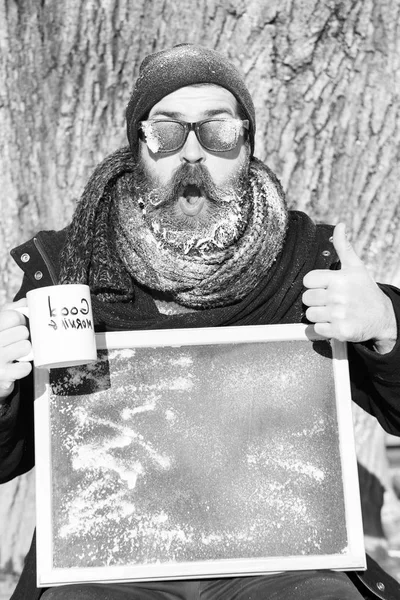 Bonito homem feliz, hipster, com barba e bigode em óculos escuros cobertos com geada branca segura placa preta em branco, polegar para cima e xícara de café no dia de inverno no fundo natural, cópia spac — Fotografia de Stock