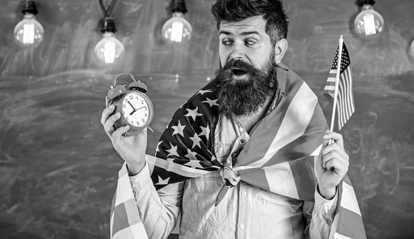 Человек с бородой на удивленном лице держит флаг США и часы, доску на заднем плане. Американский учитель с американскими флагами держит будильник. Концепция американской системы образования — стоковое фото