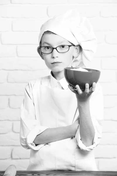 Jeune garçon petit chef cuisinier mignon en uniforme blanc et chapeau sur le visage sérieux avec des lunettes debout près de la table et tenant bol rouge sur fond de mur de briques — Photo