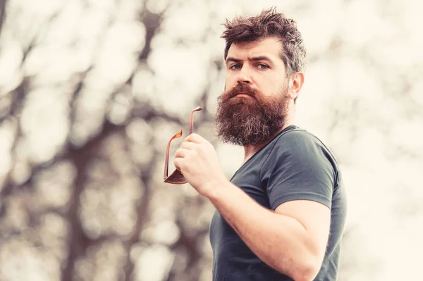 Young muscular bearded man standing in park side view, active lifestyle concept. Handsome man with blue eyes taking off his sunglasses