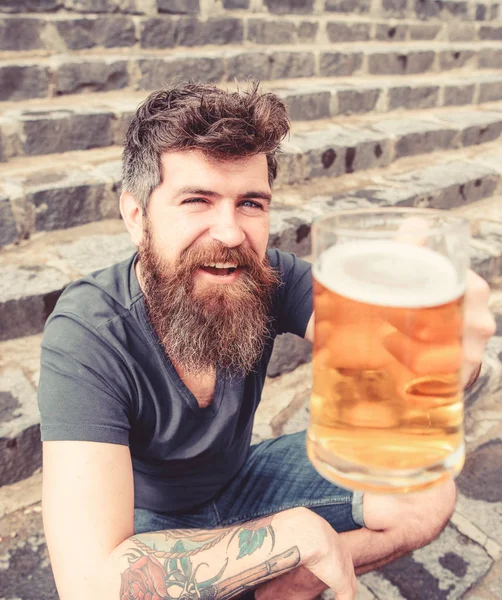 Hipster sur le visage joyeux boit de la bière en plein air. Concept de célébration. L'homme à la barbe et à la moustache tient un verre avec de la bière tout en s'asseyant sur des escaliers en pierre, déconcentré. Un type qui lève du verre avec de la bière. — Photo