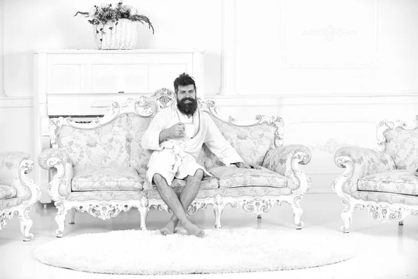 Homem com barba e bigode gosta de manhã, enquanto sentado no sofá de luxo à moda antiga. Homem alegre em roupão de banho bebe café em hotel de luxo de manhã, fundo branco. Conceito de vida de luxo — Fotografia de Stock