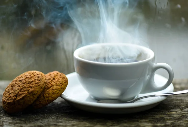 Coffee time on rainy day. Fresh brewed coffee in white cup or mug on windowsill. Wet glass window and cup of hot caffeine beverage. Coffee drink with oat cookies dessert. Enjoying coffee on rainy day