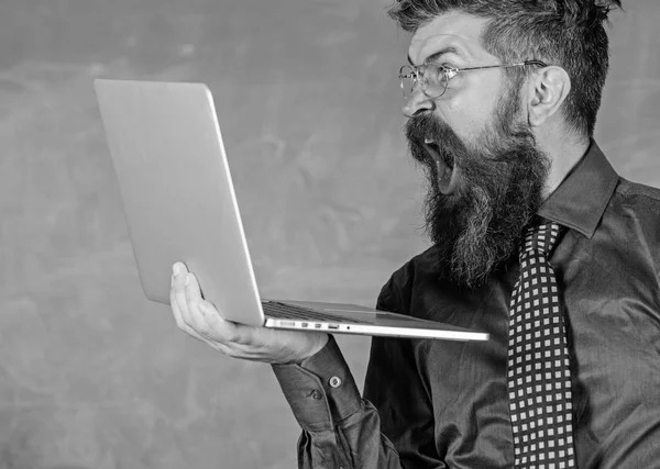 Schockierende Informationen. Hipster-Lehrer mit Brille und Krawatte hält Laptop beim Internetsurfen. Lehrer bärtigen schockierten Mann mit modernen Laptop surfen Internet-Kreidetafel Hintergrund. Schockierende Nachricht — Stockfoto