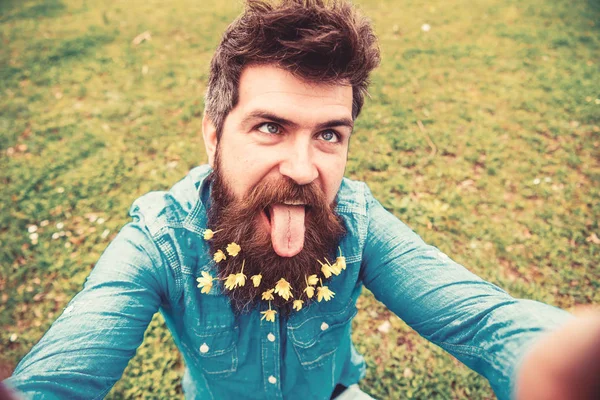 Küçük kırlangıçotu çiçeklerle selfie fotoğrafta sakallı adam. Hippi selfie alarak yüzündeki dil ile neşeli yüz buruşturma ile. Adam bahar, yeşil arka plan, defocused sahiptir. Neşeli ruh kavramı