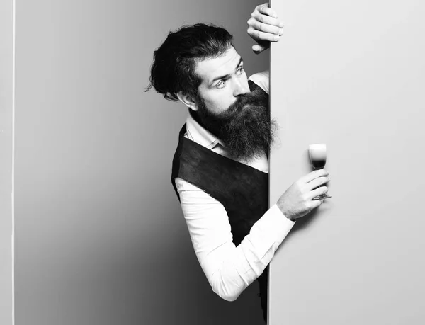 handsome bearded man with long beard and mustache has stylish hair on surprised face holding glass of alcoholic shot in vintage suede leather waistcoat on colorful studio background, copy spac