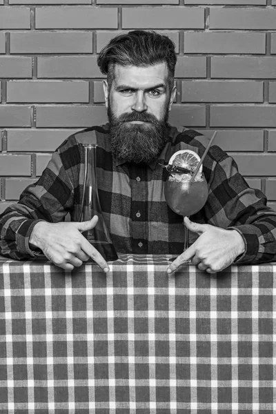 Bartender recommends to try beverage. Barman with beard on strict face pointing down with index finger. Man in checkered shirt near bottle and cocktail, brick wall background. Bar menu concept