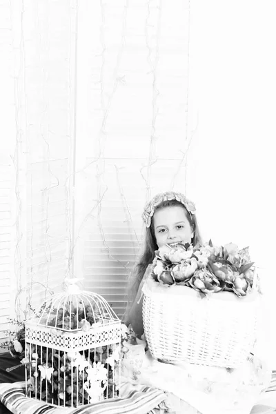 Jovem sorrindo menina com grinalda no cabelo encaracolado e vestido bonito sentado com cestas de peônias decorativas rosa e gaiola com flores no fundo do estúdio branco, cópia spac — Fotografia de Stock