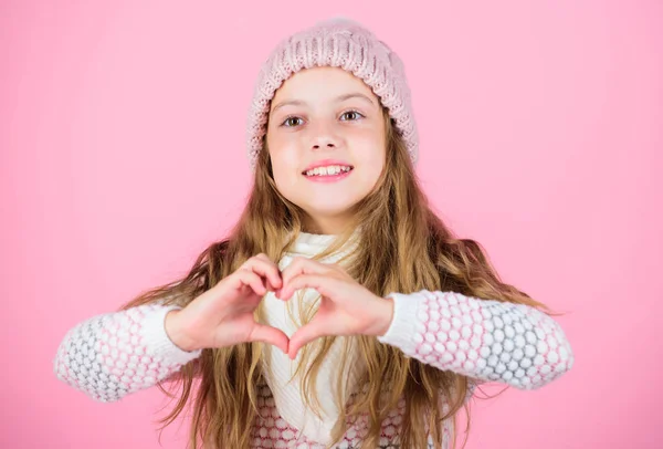 Chica usar sombrero de punto fondo rosa. Cuida el cuero cabelludo y el cabello este invierno. Prevenir daños en el cabello de invierno. Consejos para el cuidado del cabello de invierno que debe seguir. Niño pelo largo sonriente mostrar gesto del corazón — Foto de Stock