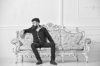 Sakallı ve bıyıklı lüks oturma odasında boş zaman harcıyor. Hipster düşünceli yüzünde yalnız oturuyor. Zengin ve yalnız kavramı. Lüks daire sahibi beyaz duvar arka koltukta oturur