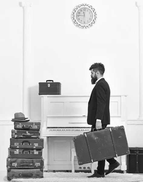 Butler and service concept. Macho attractive, elegant carries vintage suitcases, side view. Man with beard and mustache wearing classic suit delivers luggage, luxury white interior background