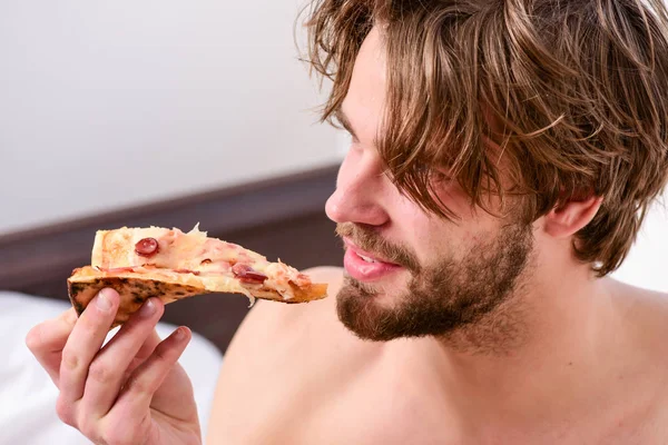 Servizio di consegna cibo. L'uomo sexy mangia pizza sdraiato sul letto. Giovane che riposa a casa con un nudo e una pizza. Mangiare pizza a letto . — Foto Stock