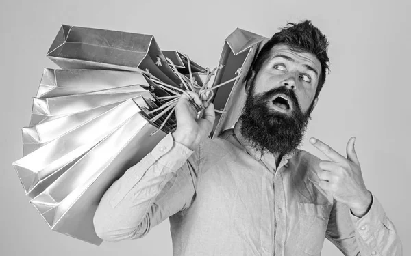 Concepto de venta y descuento. Un tipo de compras en temporada de ventas, apuntando a las bolsas. Hipster en adicto a la cara sorprendido shopaholic. Hombre con barba y bigote lleva montón de bolsas de compras, fondo gris —  Fotos de Stock