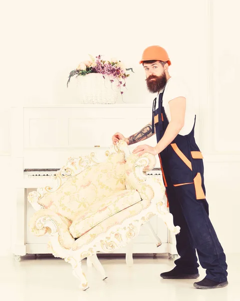 Relocating concept. Loader moves armchair for move out. Courier delivers furniture in case of move out, relocation. Man with beard, worker in overalls and helmet lifts up armchair, white background