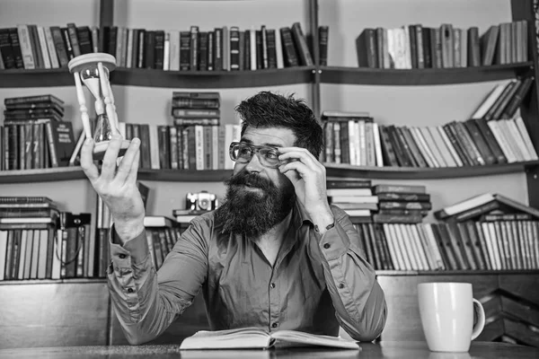 Profesor o estudiante con barba estudiando en la biblioteca. Científico con anteojos se sienta en la mesa y mira reloj de arena. Hombre ocupado leyendo libros, estanterías en el fondo. Concepto de lección y tiempo —  Fotos de Stock