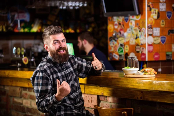 High calorie snack. Hipster relaxing at pub. Pub is relaxing place to have drink and relax. Brutal hipster bearded man sit at bar counter. Man with beard drink beer eat burger menu. Enjoy meal in pub