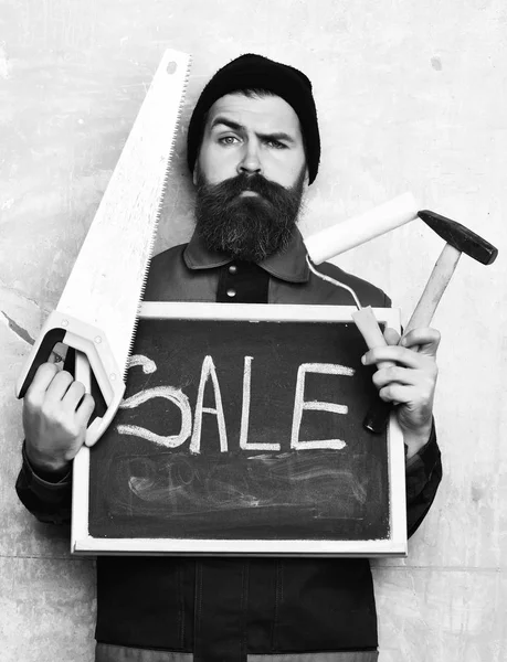 Bearded man holding various building tools and board, serious face — Stock Photo, Image