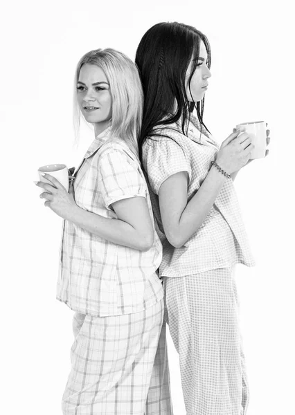 Glada flickor i pyjamas med en muggar kaffe, isolerad på en vit bakgrund. Morgon kaffe konceptet. Systrarna stå rygg mot rygg och dricka kaffe — Stockfoto