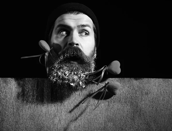 handsome bearded man or guy in winter hat with fashionable mustache on surprised face and snow in beard with decorative valentines hearts on sticks near red paper on black background, copy spac