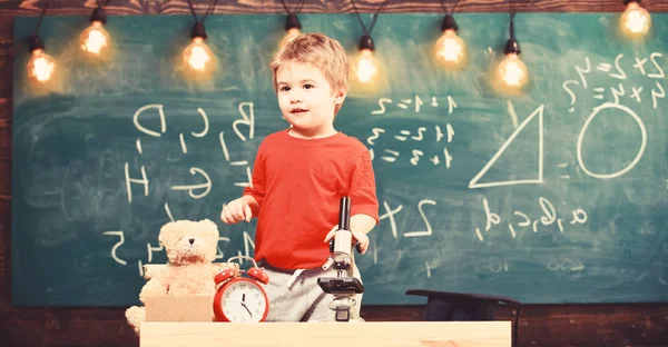 Kind, Pupille mit ruhigem Gesicht unter dem Mikroskop. begeistertes Schüler-Konzept. Junge neben Mikroskop im Klassenzimmer, Kreidetafel im Hintergrund. Erste Ehemalige interessiert an Studium, Lernen, Bildung — Stockfoto