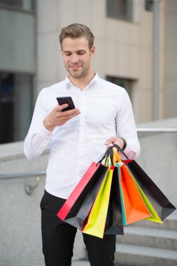 İşadamı kullanımı alışveriş uygulaması. Adam alışveriş torbaları tutun telefon kentsel arka planı süre taşır. Alışveriş online başarılı bir iş adamı. Meşgul insanlar online hizmet için teşekkür ederiz. Online alışveriş