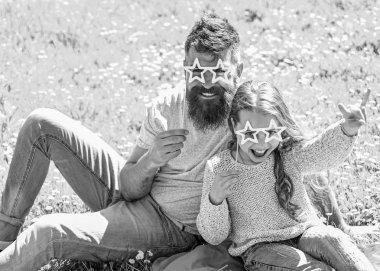 Father and daughter sits on grass at grassplot, green background. Superstar concept. Child and dad posing with star shaped eyeglases photo booth attribute at meadow. Family spend leisure outdoors clipart