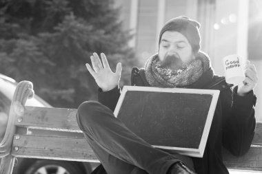 Heyecanlı adam, sakal ve bıyık beyaz frost ile kaplı sakallı hippi boş kara tahta veya chalkboard ve Kupası ile tahta bankta doğal zemin, kopya spac üzerine karlı kış gününde oturur