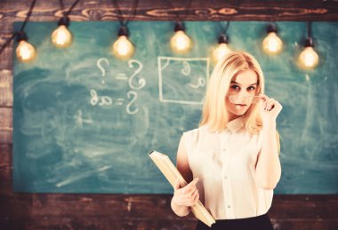 Öğretmen gözlük, kendinden emin görünüyor sınıf, arka plan kara tahta stand. Çekici öğretim görevlisi kavramı. Kadın kitap ile ders başlıyor, izleyici olarak gözlük çekerken gazes