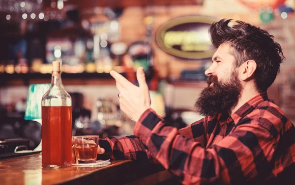 Hipster com barba pediu garrafa cheia de álcool. Conceito de relaxamento. O homem bebe uísque ou conhaque. Homem com cara feliz senta-se perto do balcão do bar. Guy passar o lazer no bar, fundo desfocado — Fotografia de Stock