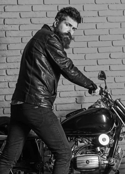 Masculine hobby concept. Man with beard, biker in leather jacket near motor bike in garage, brick wall background. Hipster, brutal biker on serious face in leather jacket sits down on motorcycle