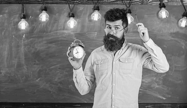 Man with beard and mustache on forgetful face itching head. Bearded hipster holds clock, chalkboard on background, copy space. Discipline concept. Teacher in eyeglasses holds alarm clock and pen — Stock Photo, Image