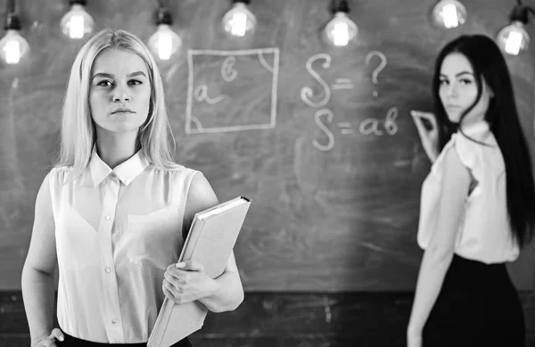 Studenten en stagiairs concept. Meisje ziet er zelfverzekerd uit terwijl dame op schoolbordachtergrond schrijft, ongeconcentreerd. Leerling, leraar in formele kleding staan in de klas. Aantrekkelijke vrouwen die zich voorbereiden op de les — Stockfoto