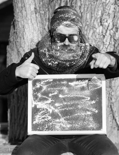 Homem sério, hipster, com barba e bigode em óculos escuros cobertos com geada branca dá polegares para cima gesto com placa preta no dia de inverno no fundo natural, cópia spac — Fotografia de Stock