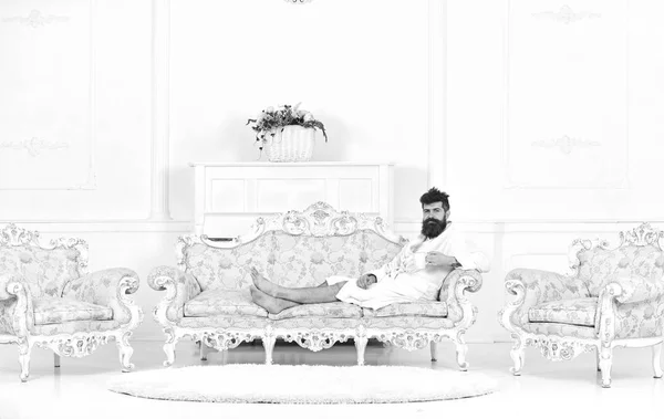 Hombre somnoliento en albornoz bebe café en hotel de lujo en la mañana, fondo blanco. Concepto de vida de lujo. Hombre con barba y bigote disfruta de la mañana sentado en un sofá de lujo a la antigua — Foto de Stock