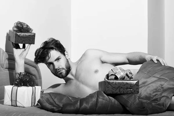 Joven guapo barbudo sexy macho hombre con barba elegante acostado desnudo en la cama bajo una manta roja con regalos o regalos de San Valentín en el fondo colorido — Foto de Stock
