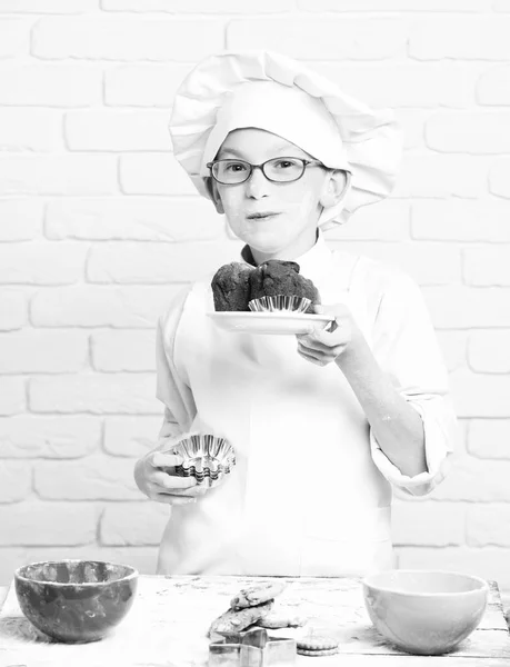 Mladý chlapec roztomilý kuchař kuchař v bílém jednotné a klobouk na mouku potřísněný obličej s brýlemi stojanu stolu s forem pro soubory cookie barevné misky a hospodářství čokoládové dorty na cihlovou zeď na pozadí — Stock fotografie