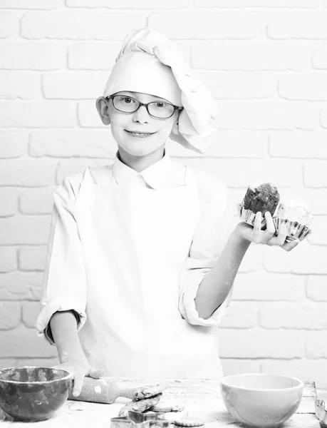 Young boy cute cook chef in white uniform and hat on stained face flour with glasses standing near table with colorful bowls and holding chocolate cakes on brick wall background — Stock Photo, Image