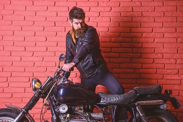 Masculine passion concept. Man with beard, biker in leather jacket near motor bike in garage, brick wall background. Hipster, brutal biker on serious face in leather jacket gets on motorcycle